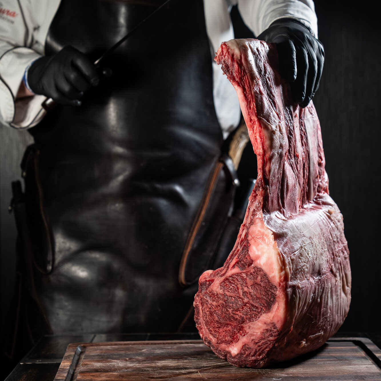 A chef in a leather apron holds a whole Dry-aged raw bone-in tomahawk rib. They are all from the station's own herd, prepared in-house.
