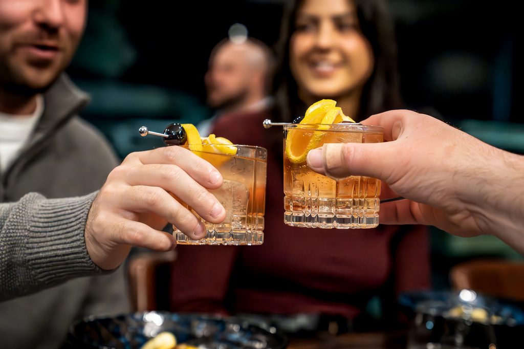 Two hands raise cocktail glasses garnished with lemon peel and a cherry, guests behind