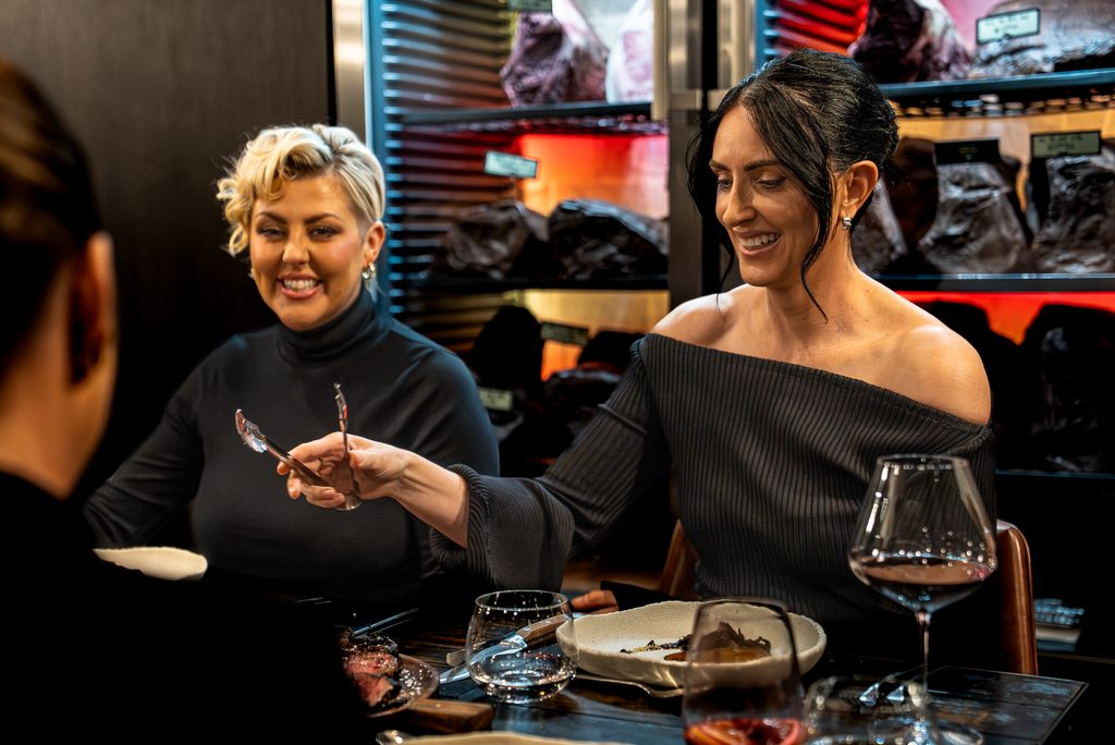 Two women laughing at the counter in front of the dry-ageing cabinet, a third guest in the foreground.