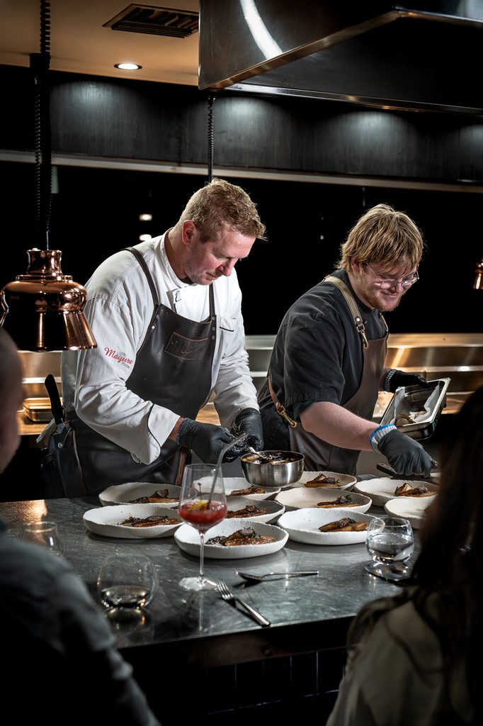 Two chefs plating dishes at the kitchen pass under copper lamps.