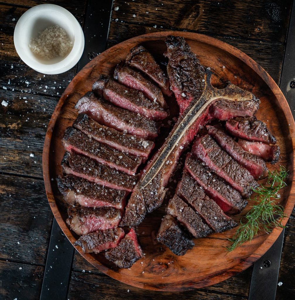 A carved T-bone steak on a round wooden platter, with a small dish of salt flakes and a sprig of herb.